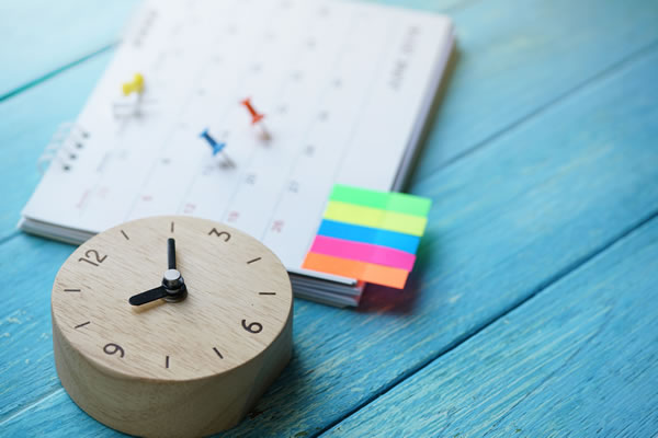 a small wooden clock on a blue table with a paper calendar with sticky notes